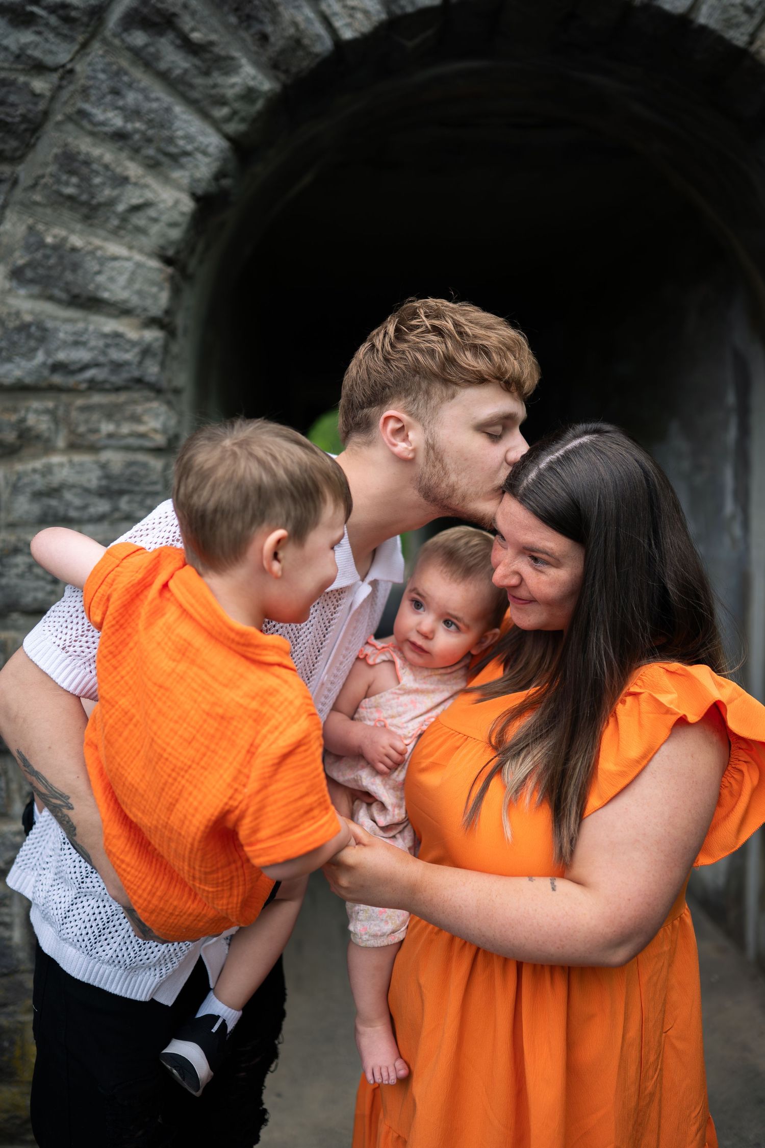 A young family in matching orange outfits shares a tender moment, with dad kissing mom on the cheek while holding two young children.