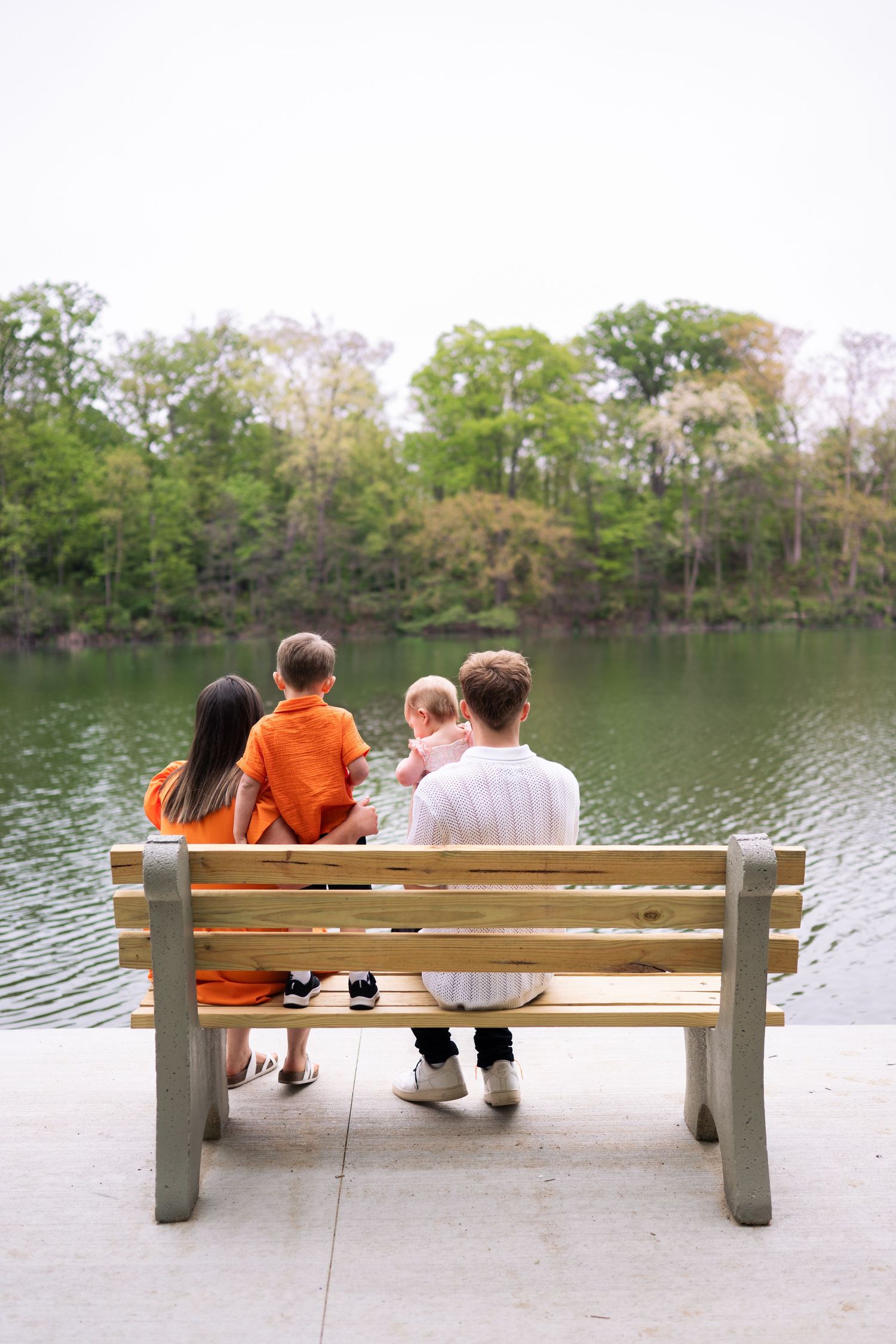 Family of four sitting on a wooden bench by a serene lake, surrounded by lush green trees, viewed from behind.