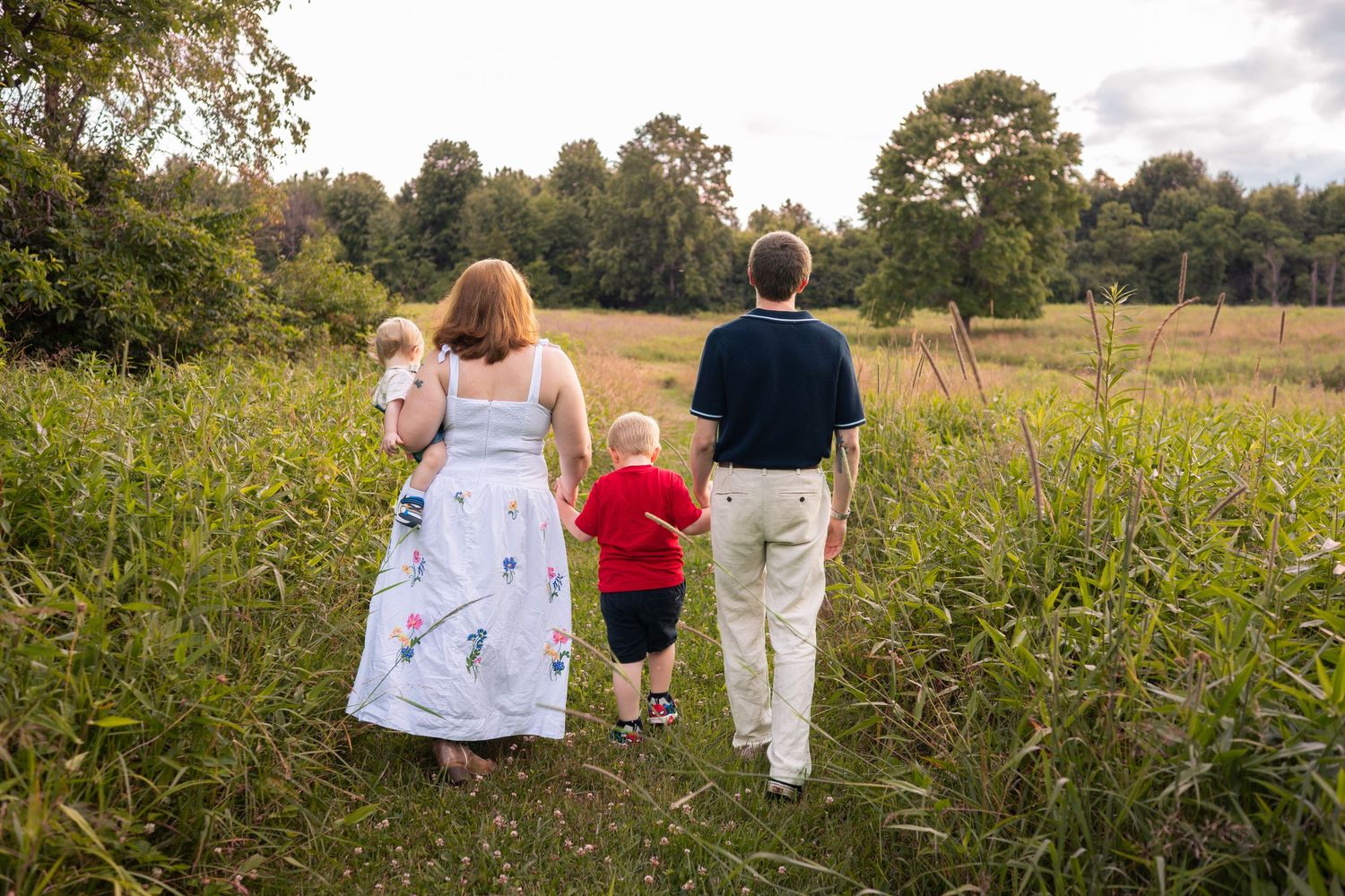 Family of four walking through a lush green meadow path, parents holding children's hands at sunset.