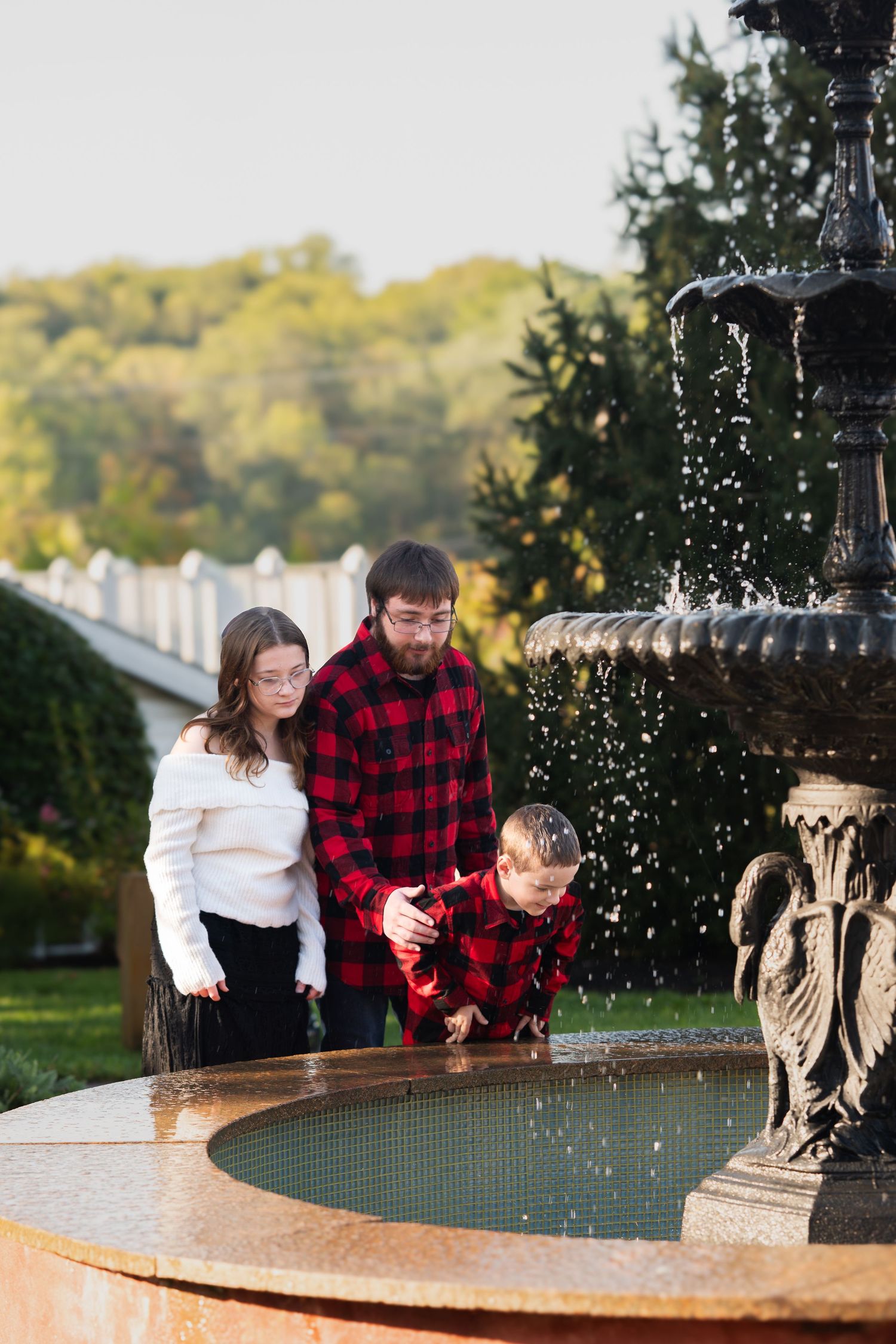 Family in matching red buffalo plaid enjoying a moment together by an ornate garden fountain on an autumn day.