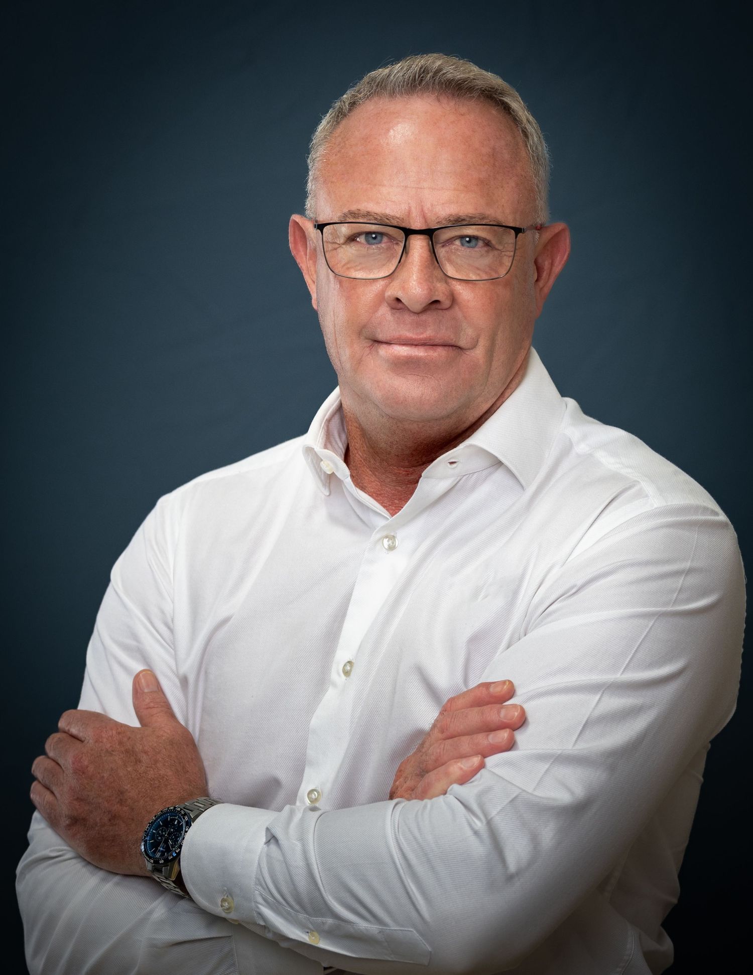 Professional headshot of a confident person wearing glasses and a white button-down shirt against a dark background.