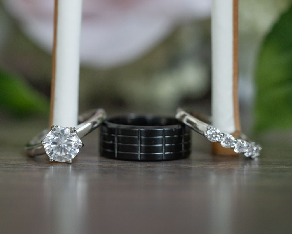 Close-up of three wedding rings arranged together, featuring two diamond bands and one black textured band on wooden surface.