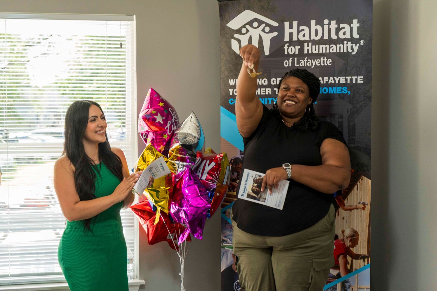 Two individuals celebrate joyfully with colorful balloons in front of a Habitat for Humanity banner.