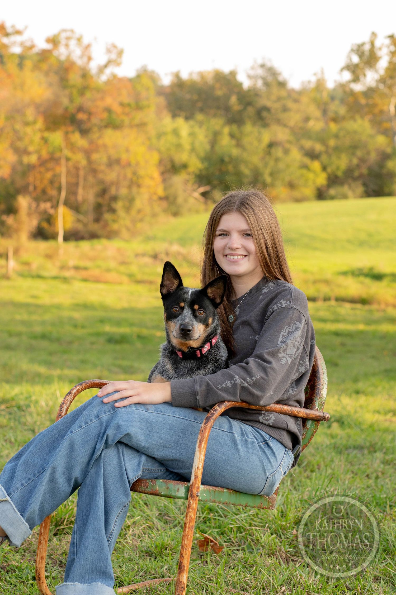 Person sits in vintage metal chair with black and white Australian Cattle Dog in sunny field.