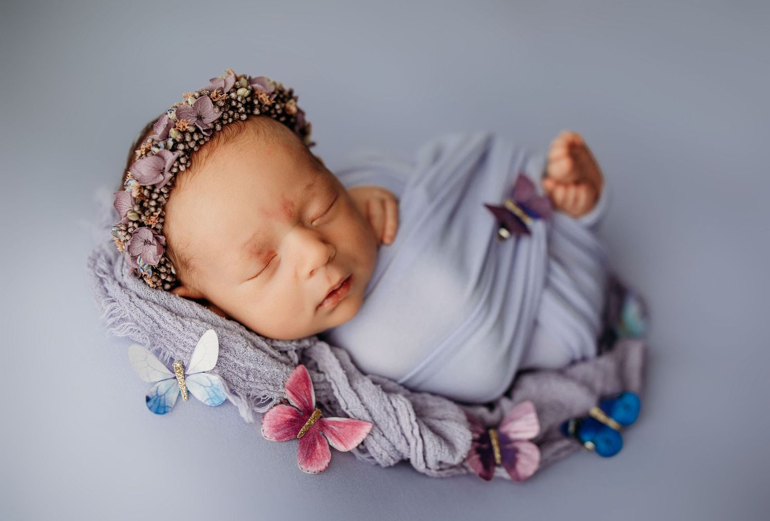 Sleeping newborn wearing a floral crown and lavender wrap on grey backdrop.