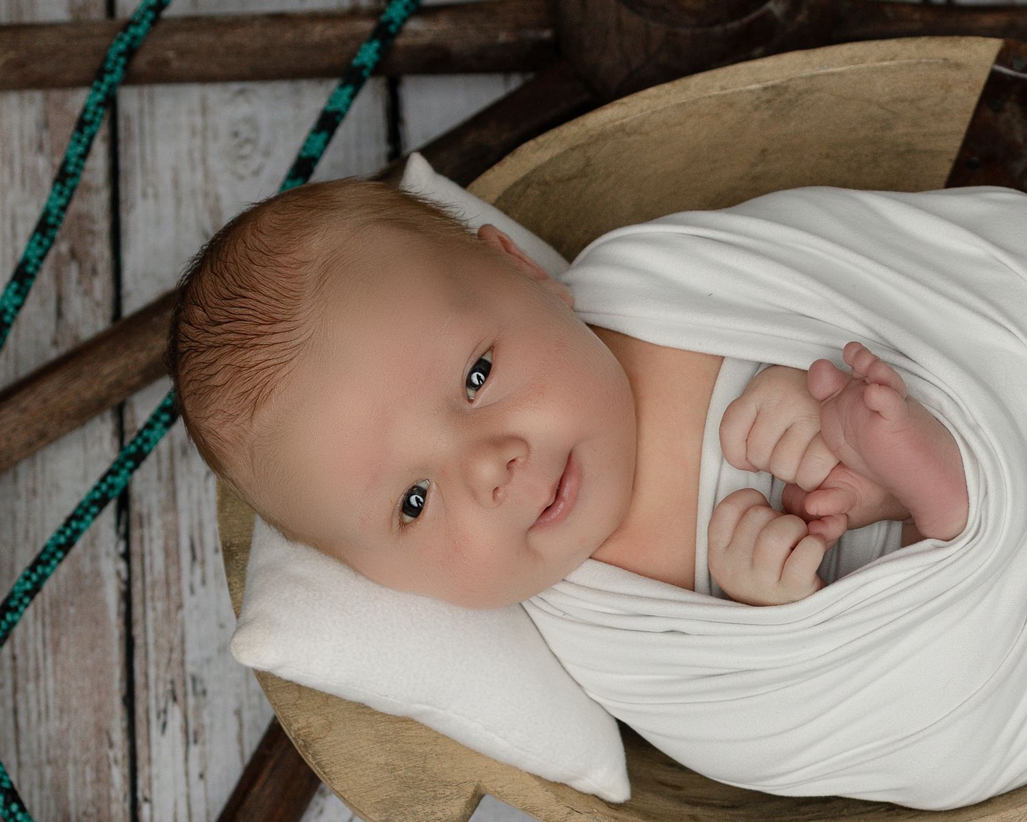 A swaddled baby lies on cozy blankets near turquoise rope against a rustic wooden backdrop.