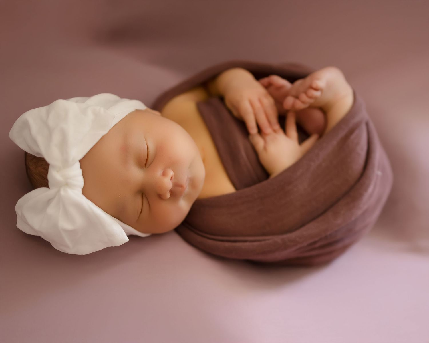 A sleeping baby swaddled in brown fabric wearing a white bow headband rests peacefully.