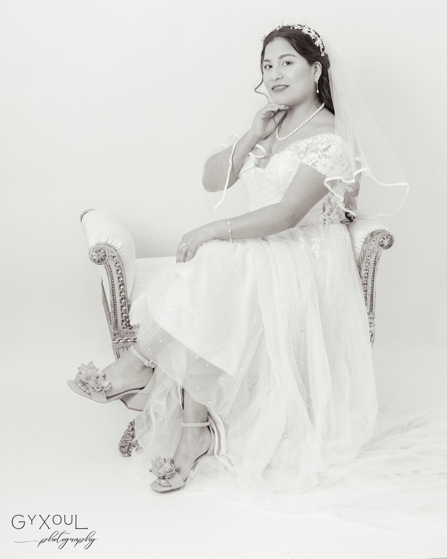 A classic bridal portrait in black and white showing full wedding attire while seated in an ornate chair.