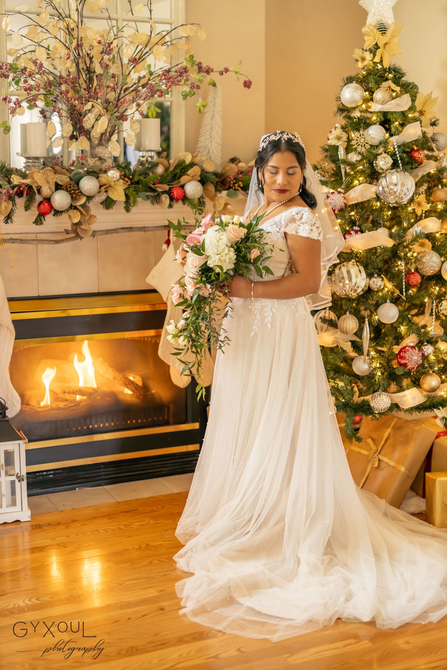 A bride stands by fireplace with Christmas decor holding cascading white and green bouquet.