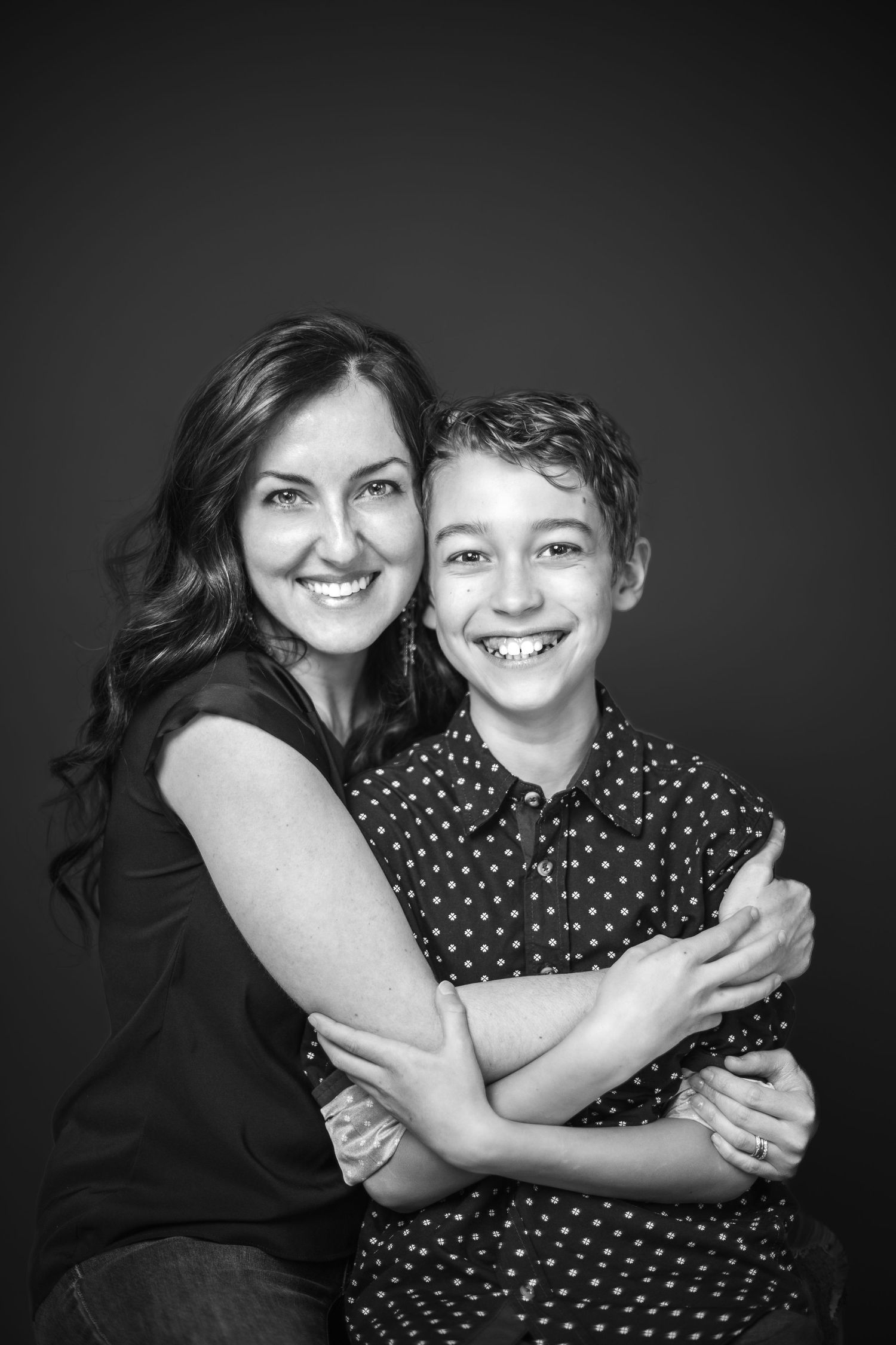 Parent and child in matching black outfits share a joyful hug while posing against a dark backdrop in monochrome.