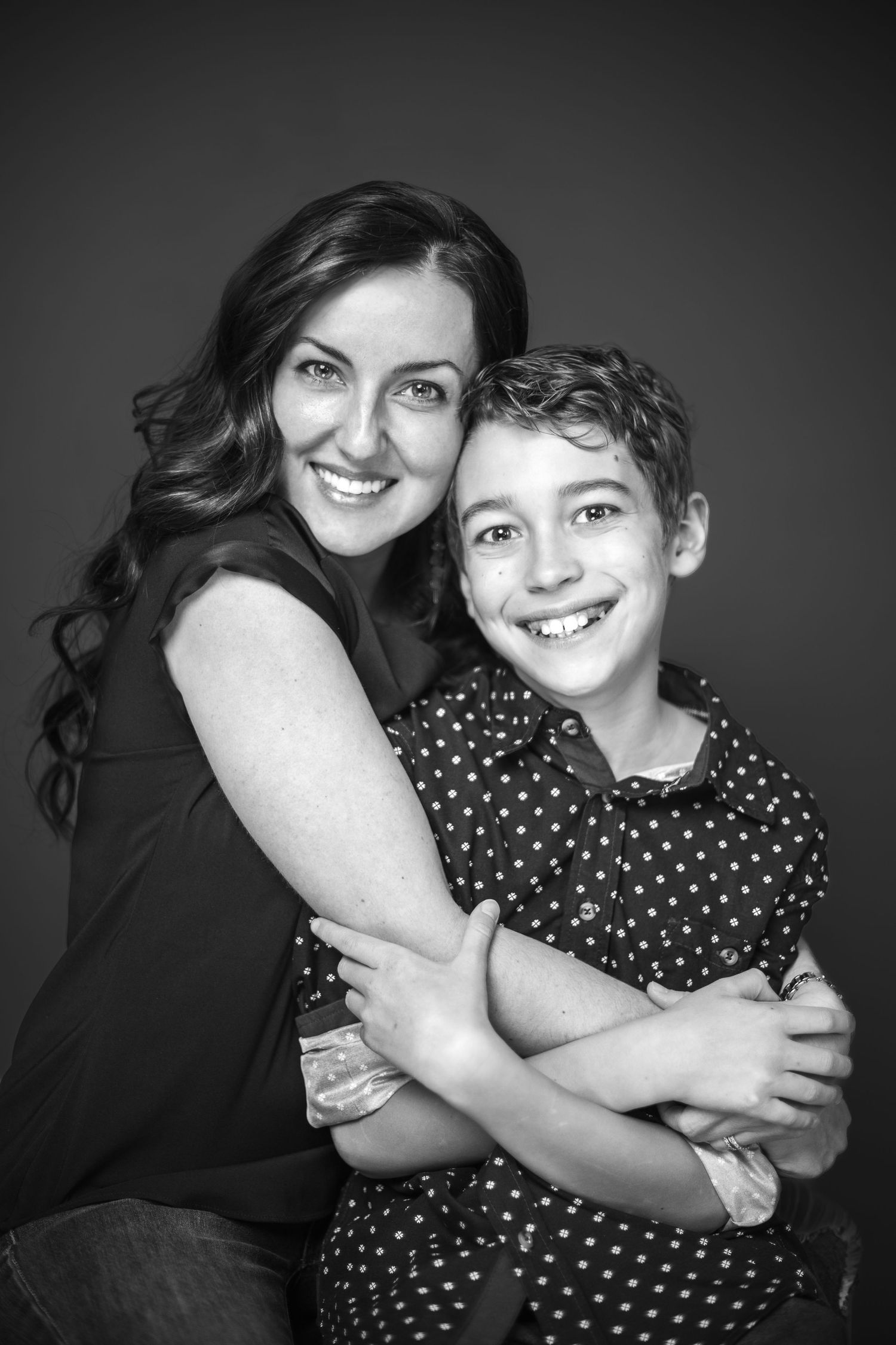Black and white portrait of a parent and child embracing and smiling at the camera against a dark background.