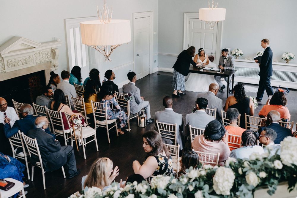 Wedding guests seated at elegant white chairs during an indoor reception at Morden Hall with floral arrangements and chandeliers.