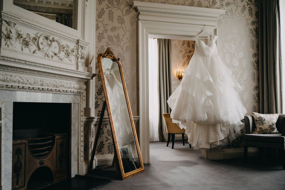 A white wedding dress hangs in an elegant room of Morden Hall with ornate moldings, vintage fireplace and gold-framed mirror.