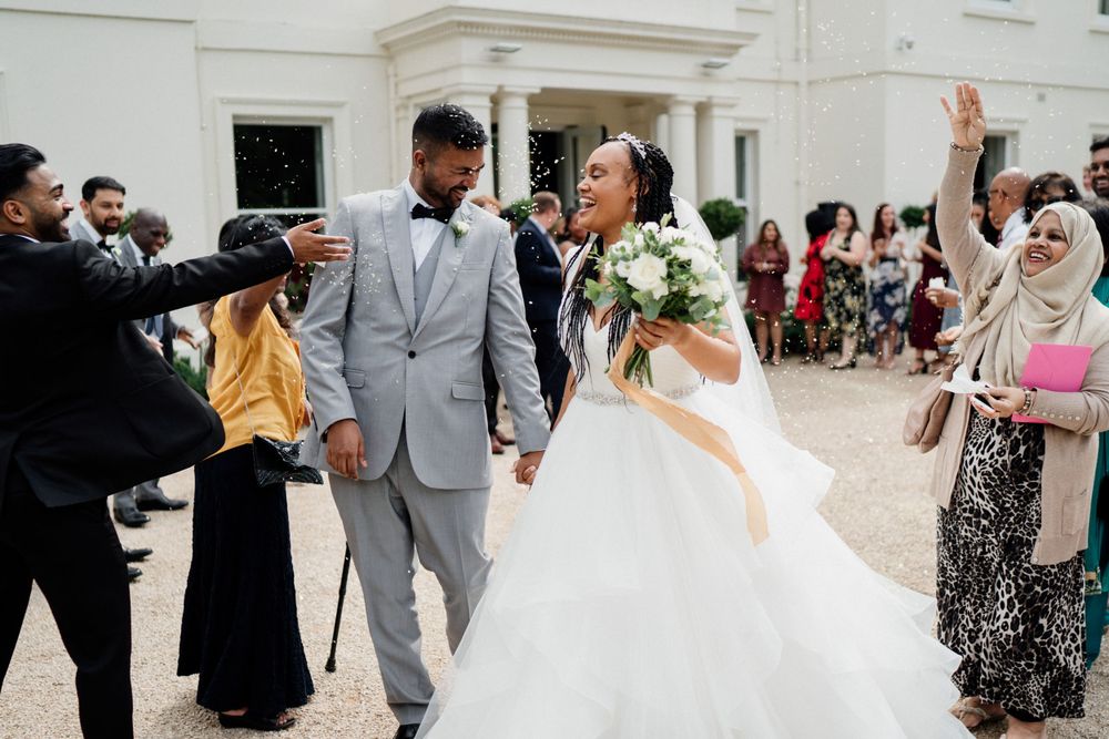 Joyful wedding celebration outside white building with couple walking while guests cheer and throw confetti.