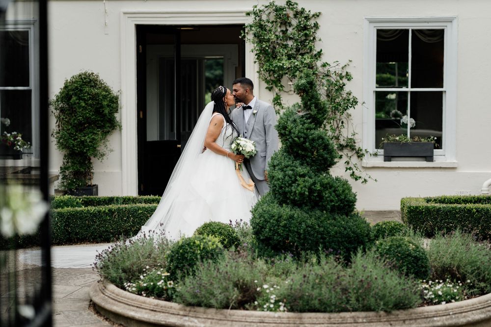 A couple in wedding attire sharing a moment in a garden with manicured hedges and climbing vines against a white building.