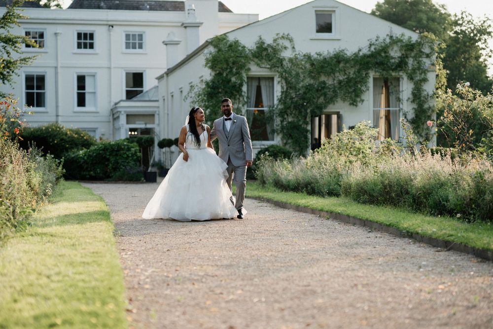 Newlyweds walk together on a gravel path beside a white mansion and garden during golden hour.