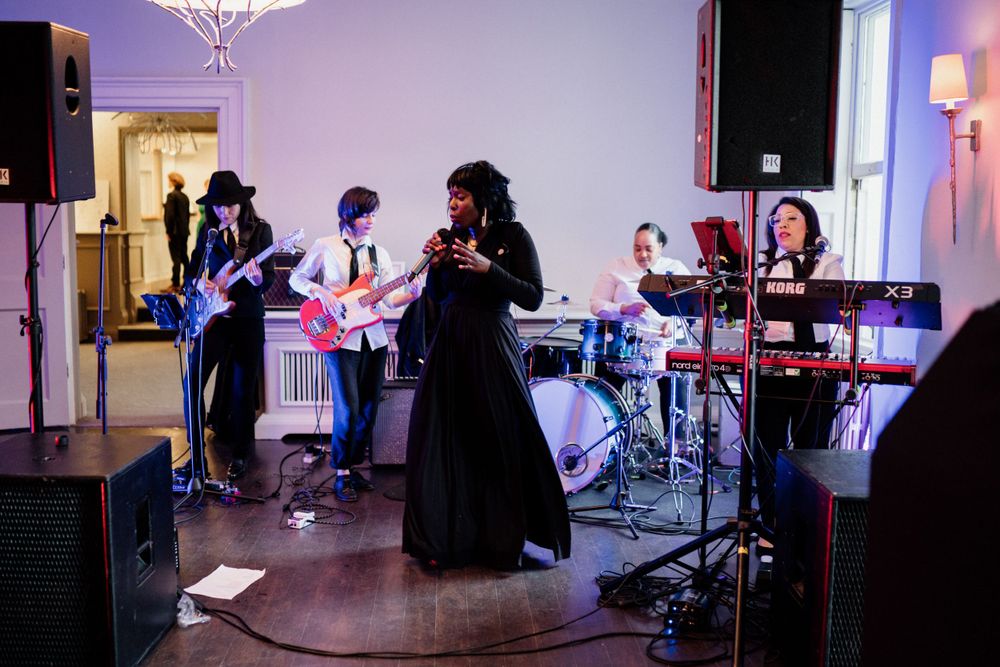 Live band performs on stage at an indoor venue with purple lighting and a singer in a black dress at the microphone.