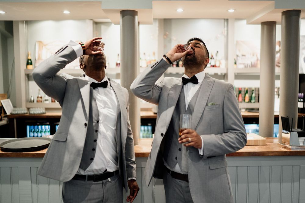Two groomsmen in matching light gray suits and bowties take shots at a wedding reception bar.