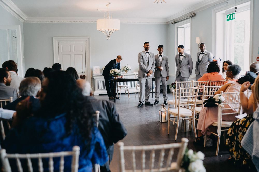 Wedding ceremony at Morden Hall guests sit in white chairs watching groomsmen at the altar in a bright modern venue.