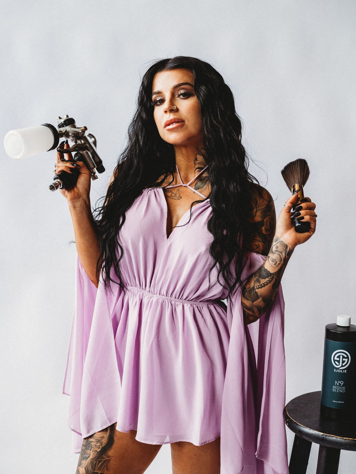 Model in lilac dress holding spray tan equipment against white backdrop.