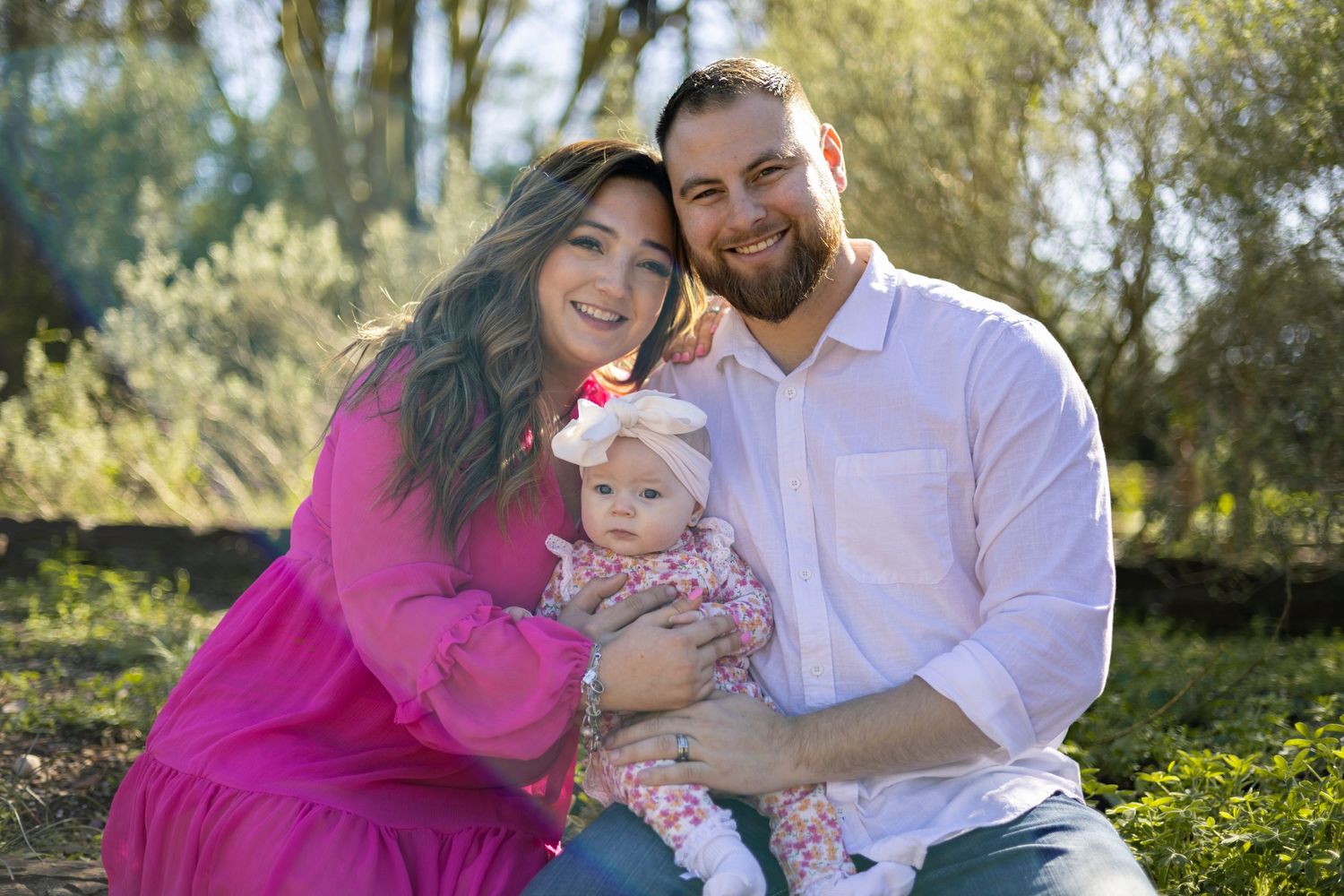Family portrait outdoors in a natural setting with vibrant pink dress and white shirt surrounded by greenery.