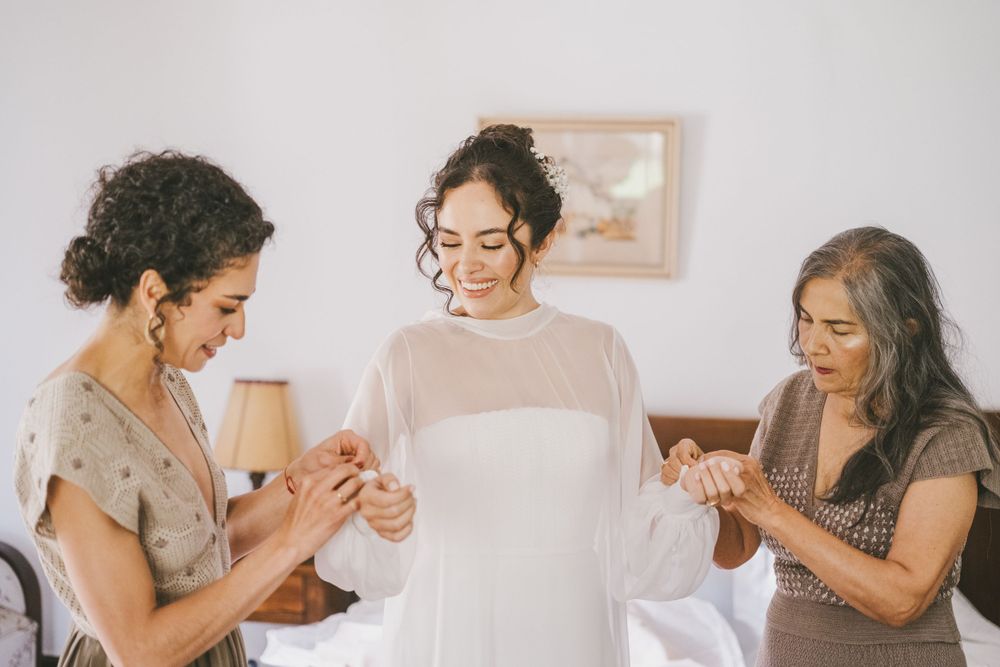 Three people helping with bridal preparations before a wedding, holding a white wedding dress in a light-filled room.