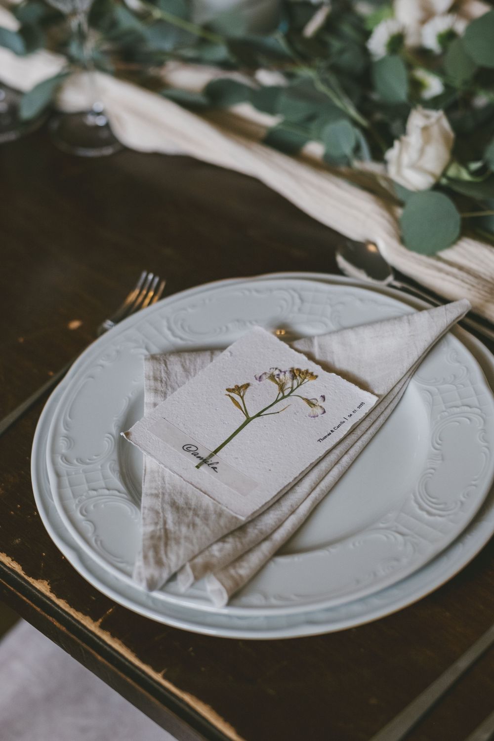 Elegant white plate with embossed details set with linen napkin and dried flowers on dark surface.