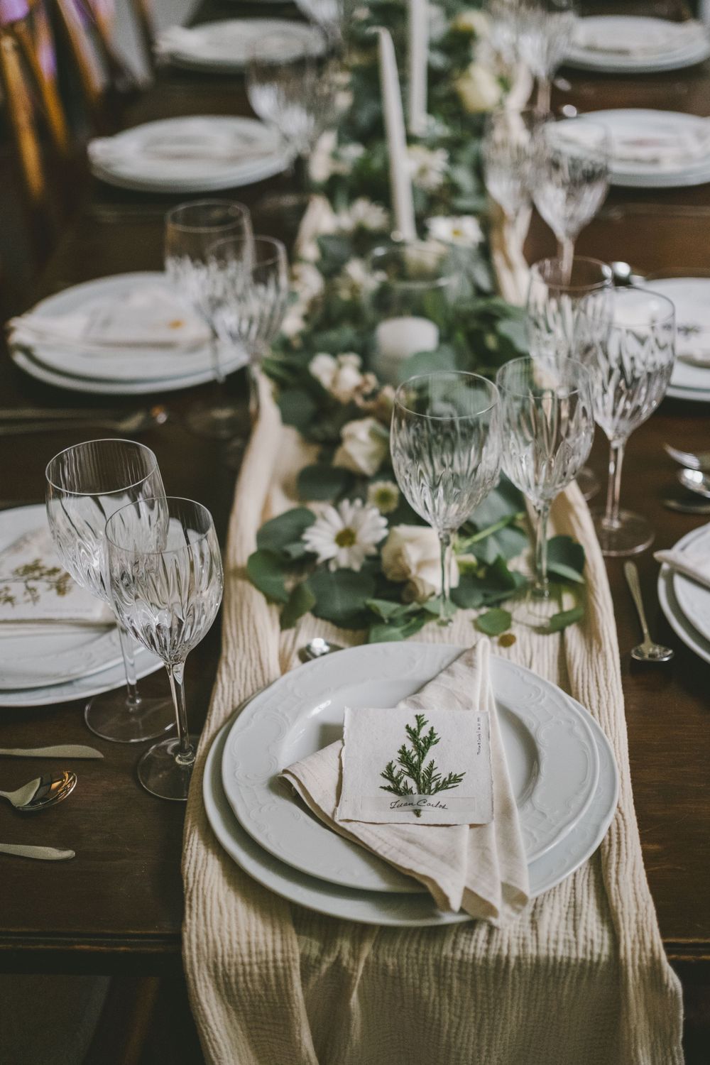 Elegant table setting with crystal glasses, white plates, and floral decor on a beige runner.