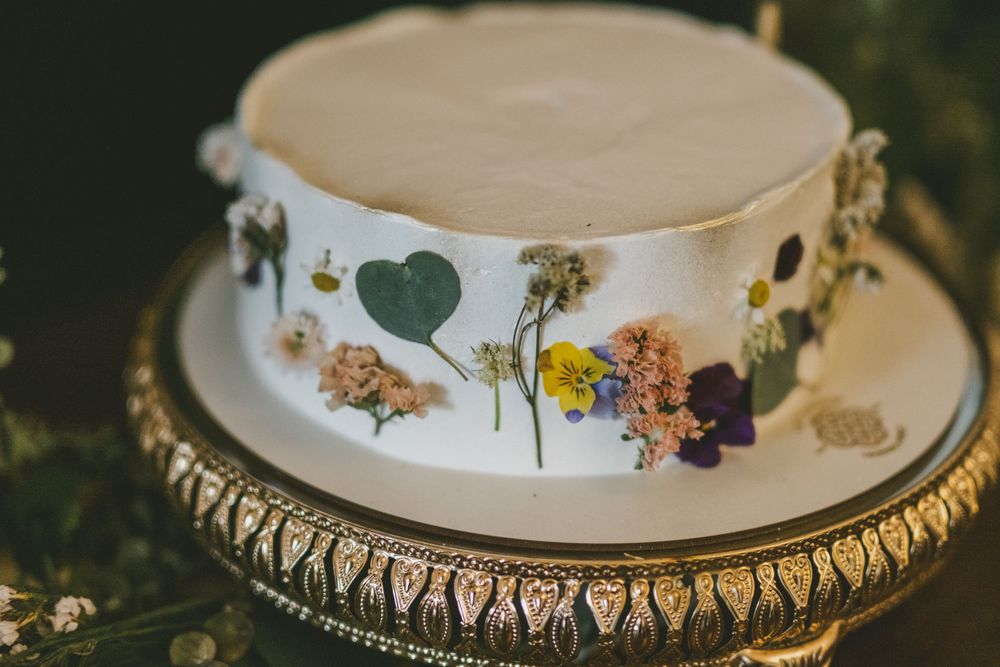 Wedding cake decorated with pressed dried flowers and hearts on an ornate gold stand.