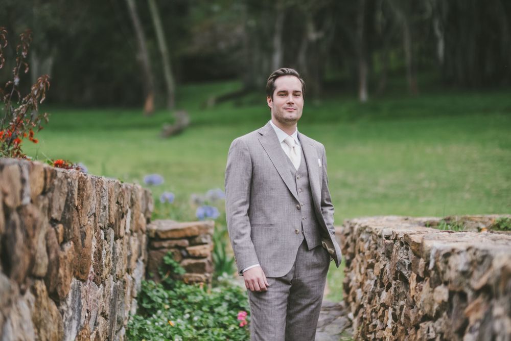 A person wearing a light gray suit stands between two rustic stone walls in a green outdoor garden setting.