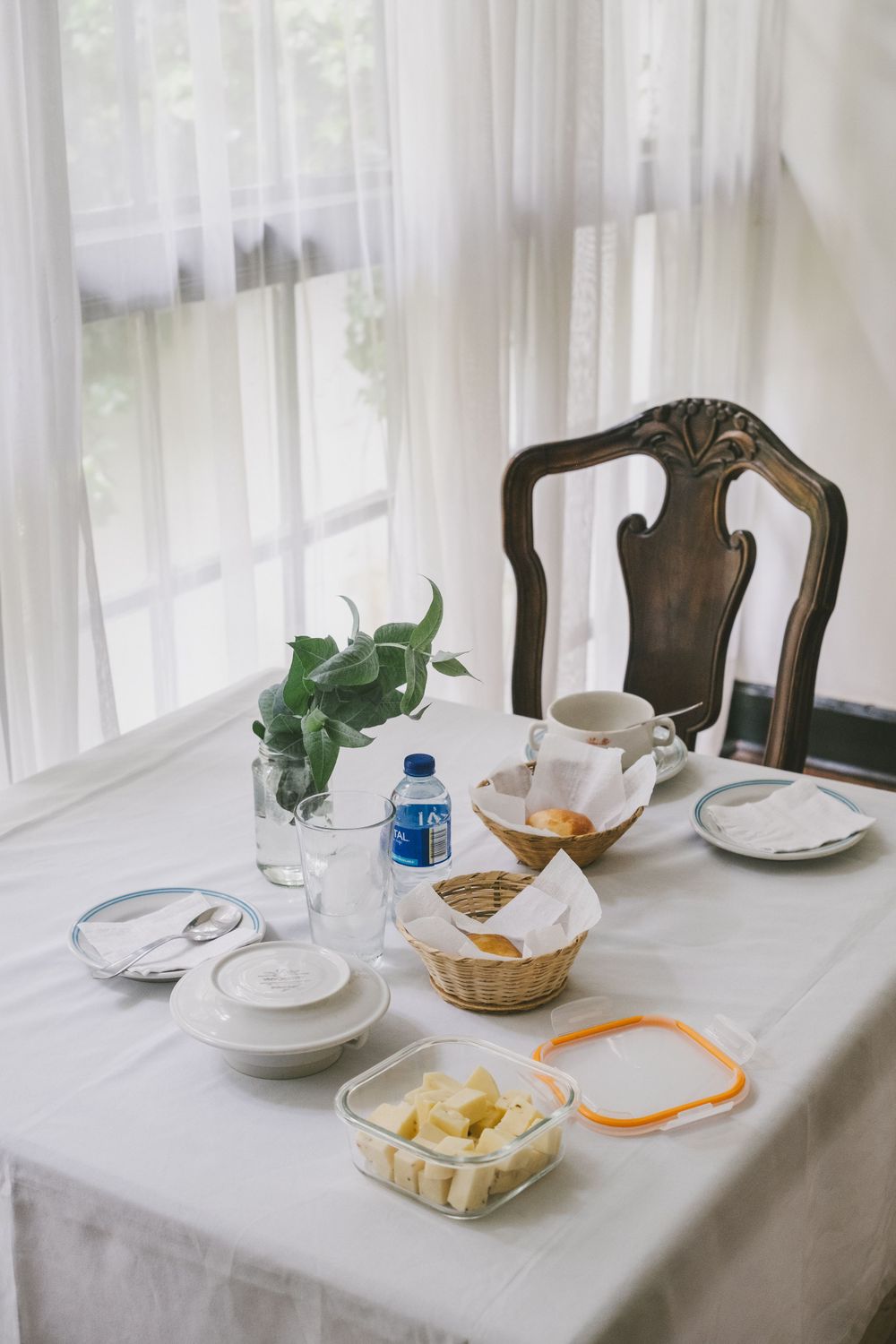 A bright breakfast table setting with white linens, plates, a water bottle, and a potted plant near sheer curtained windows.