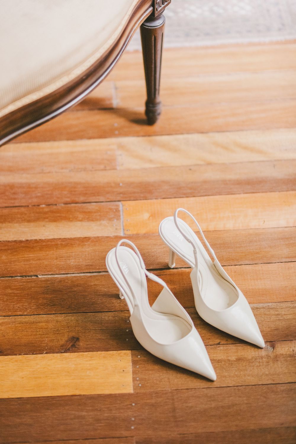 White pointed-toe slingback heels sit elegantly on a hardwood floor beside furniture.