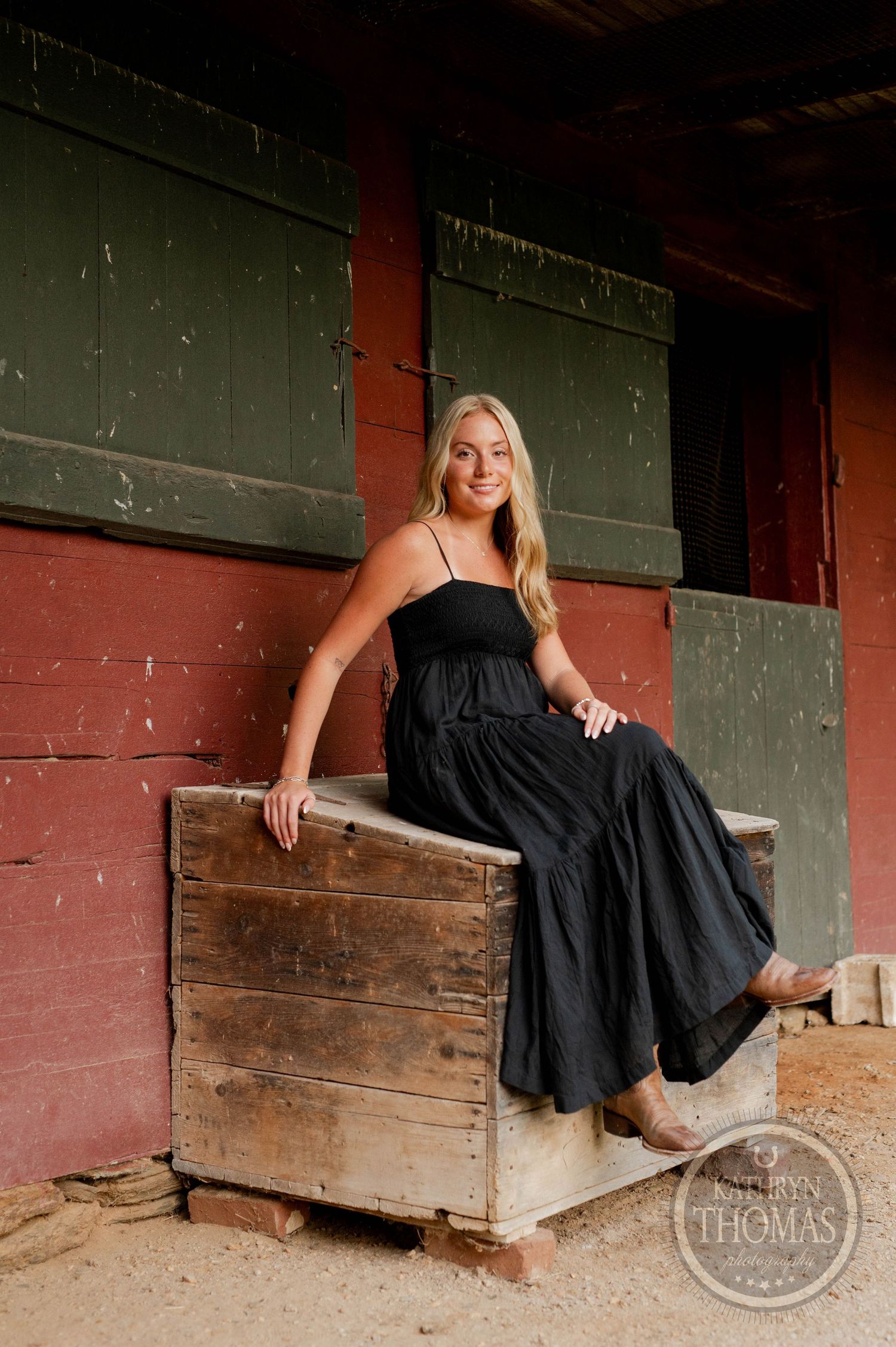 A portrait of someone in a black maxi dress sitting on a wooden crate against a rustic red barn wall.