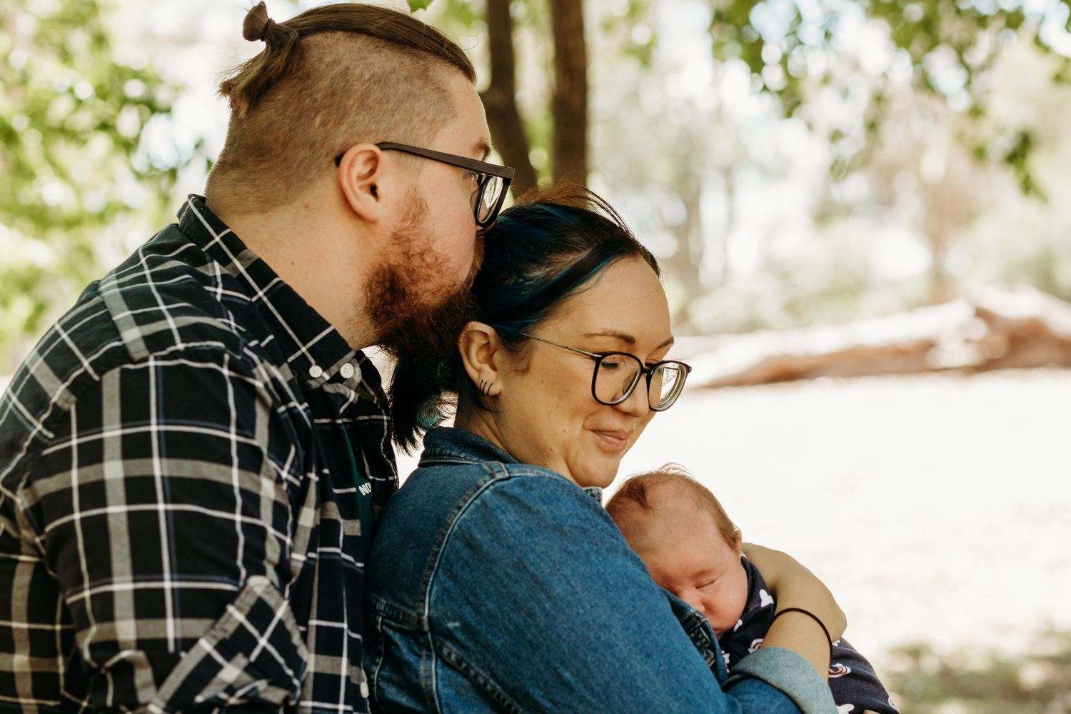 A couple wearing plaid and denim embraces their sleeping newborn baby outdoors on a sunny day.