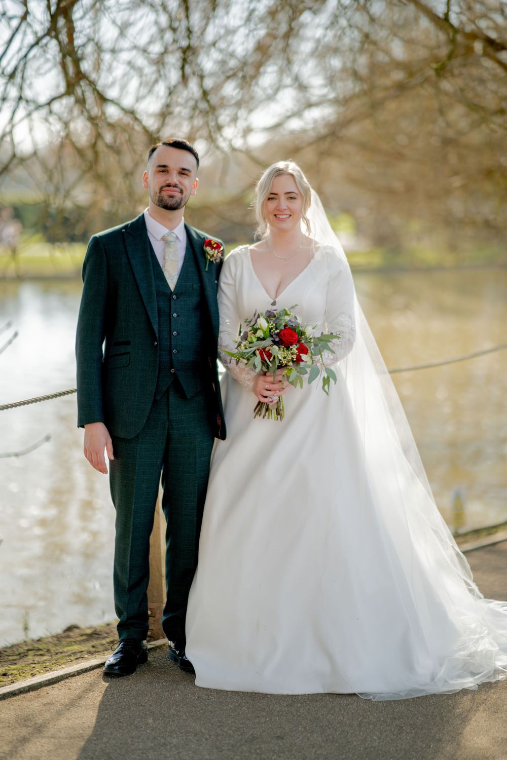 A newlywed couple poses outdoors near a lake in formal wedding attire with a bouquet of red and green florals.