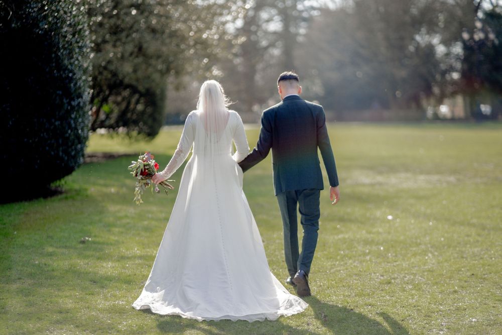 A bride and groom walk hand in hand through a sunlit garden meadow on their wedding day.