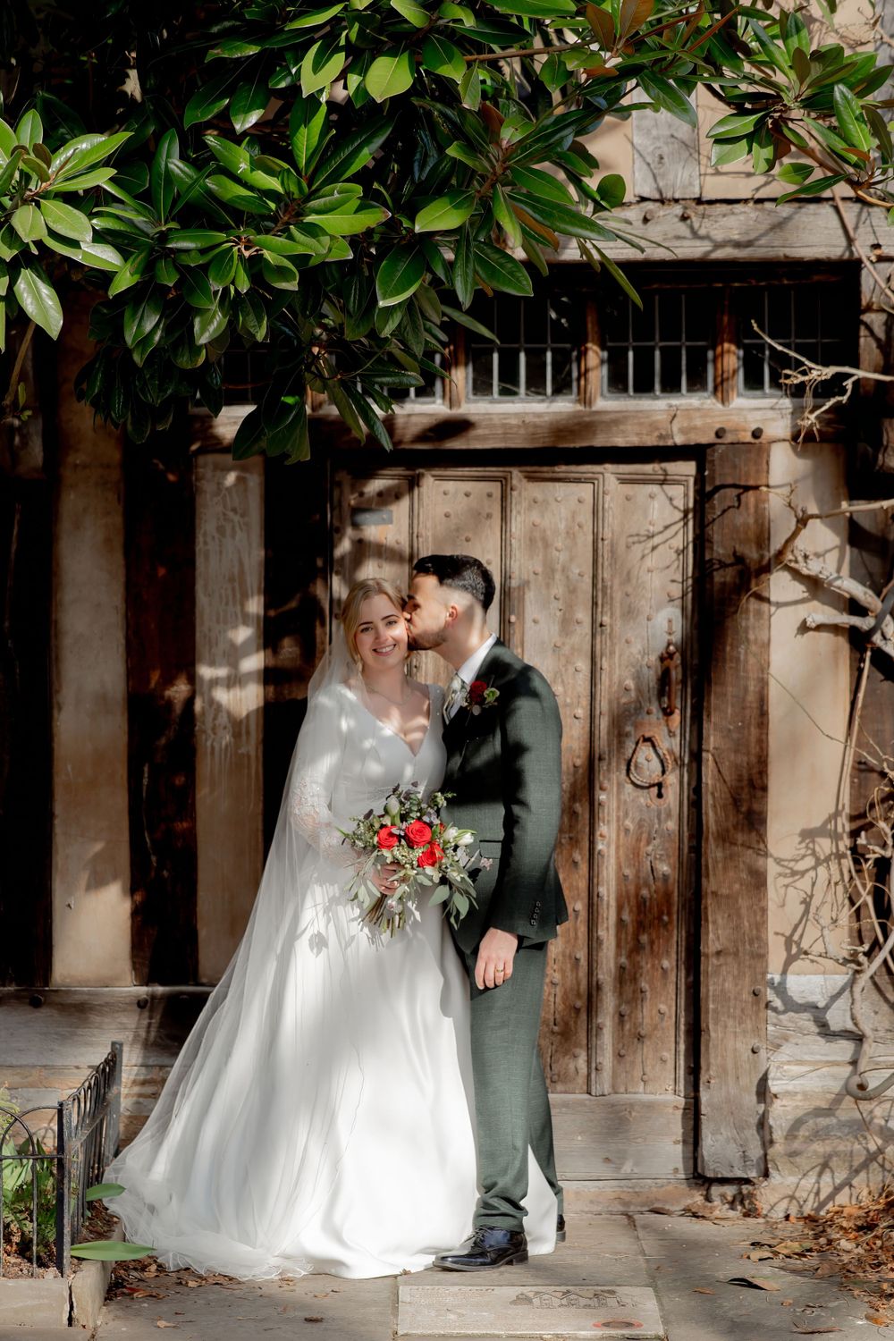 A romantic wedding portrait against a rustic wooden door framed by lush green foliage and dappled sunlight.