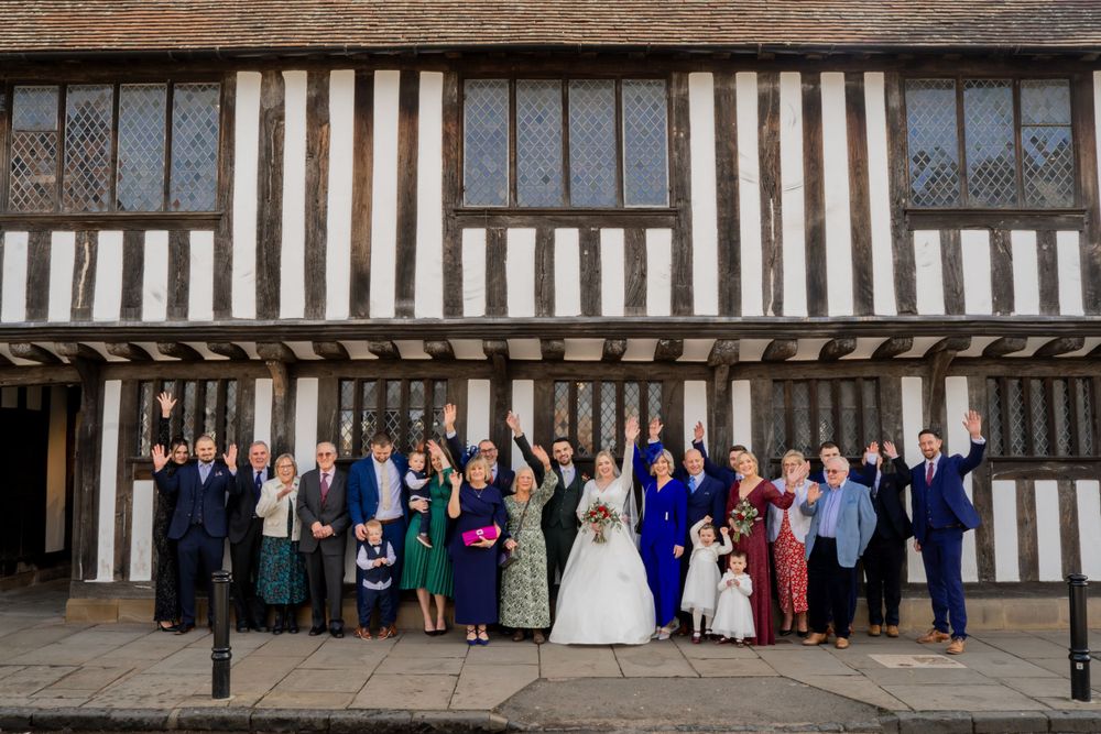 A large wedding party poses in front of a historic Tudor-style building with distinctive black and white timber framing.