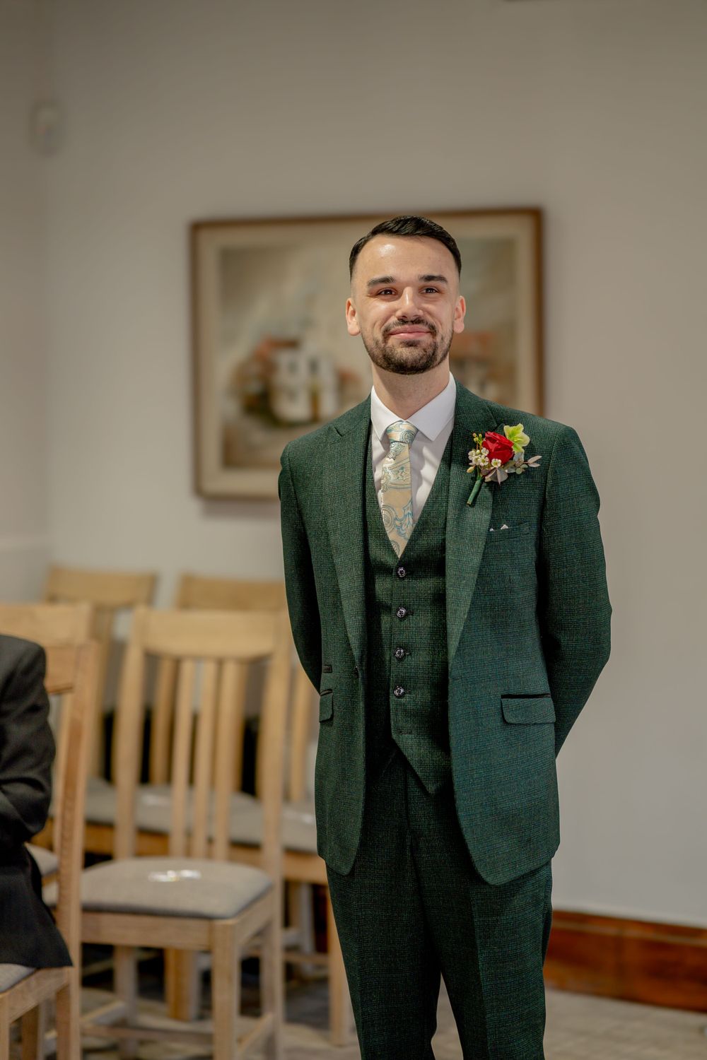 A person wearing a dark green three-piece suit with a boutonniere smiles while standing in a room with wooden chairs.