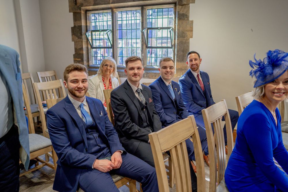 Wedding guests seated in wooden chairs wearing navy suits and bright blue attire during an indoor ceremony.