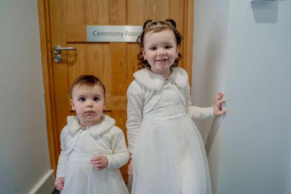 Two children in white dresses stand next to a door in a hallway.