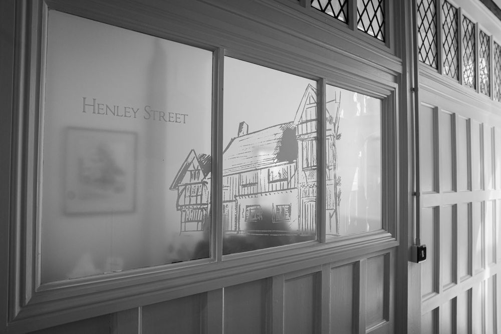 Henley Street reflected in a window pane with historic buildings visible through diamond-patterned glass.