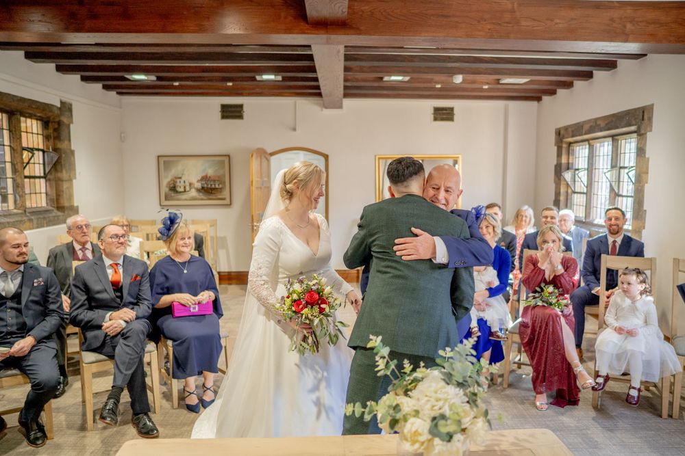 A wedding ceremony takes place in a rustic room with wooden beams, as guests look on from their seats.