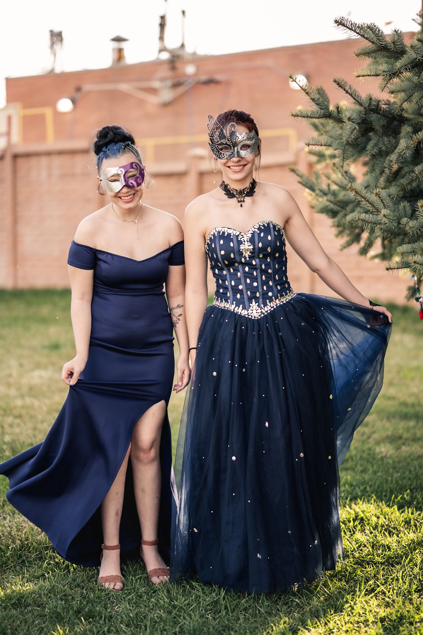 Two people in elegant navy blue formal dresses and masquerade masks pose at an outdoor event.