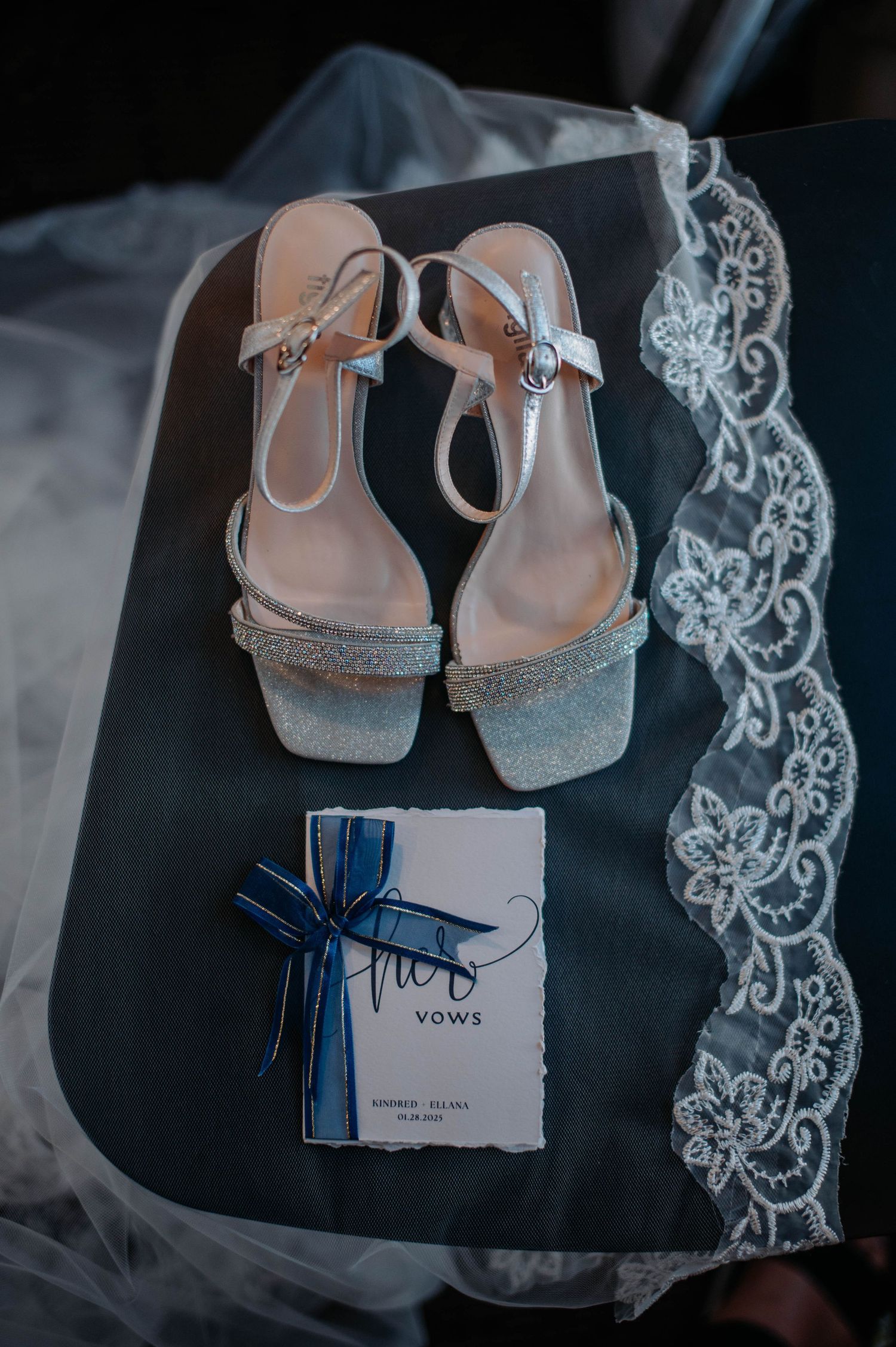 Wedding accessories including sparkly heels, lace veil and vows booklet arranged on dark surface.