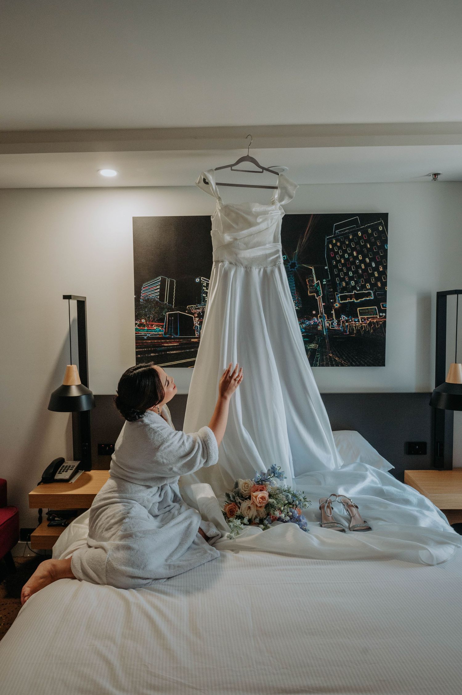 A white wedding dress hangs elegantly above a bed with city artwork in the background.