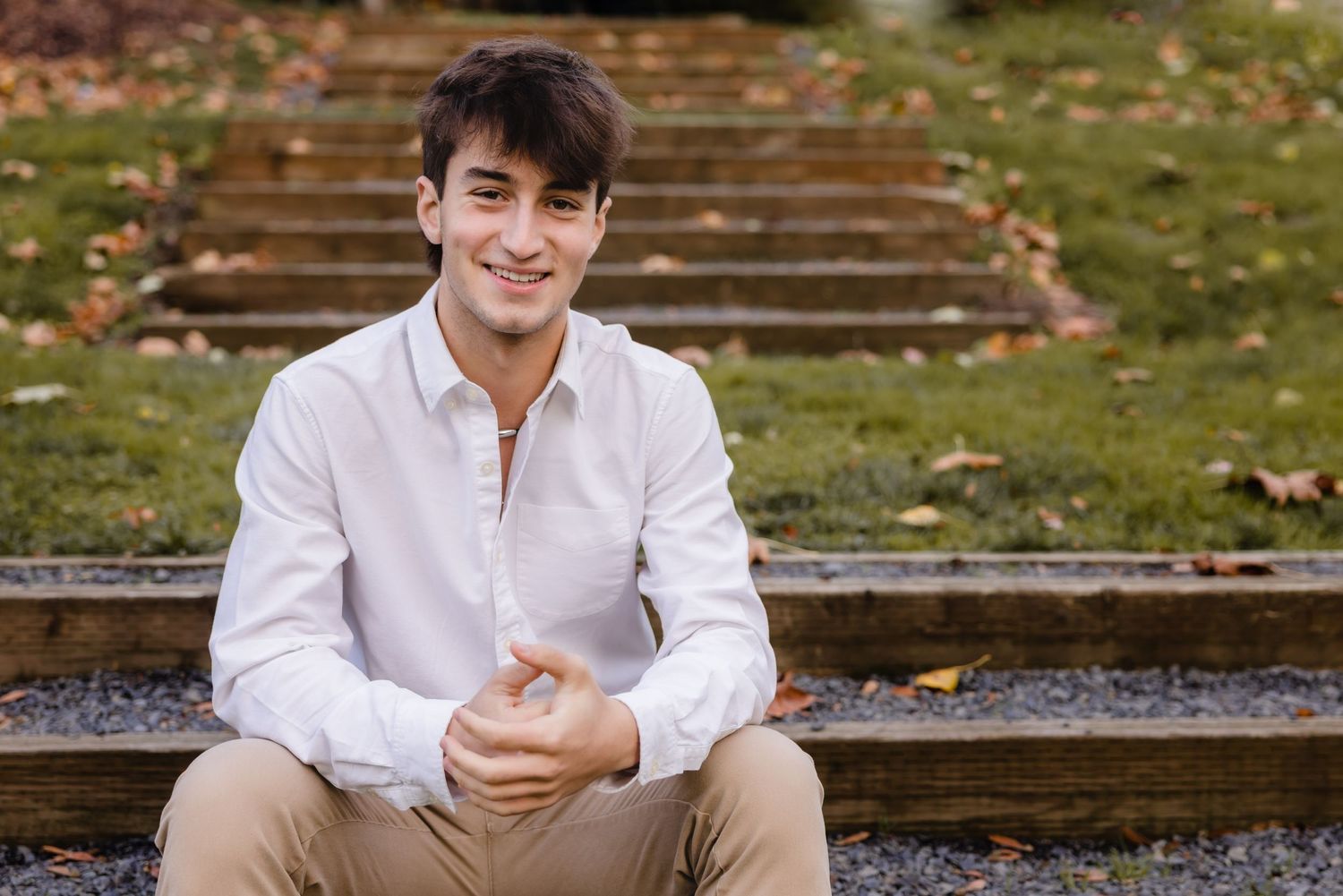 Young person in white button-down shirt sits smiling on stone steps surrounded by autumn leaves.