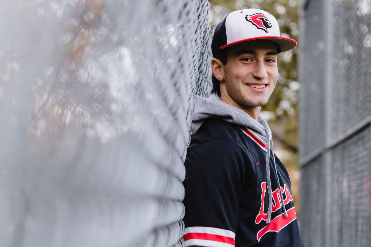 A young athlete in a Cleveland Guardians baseball jacket and cap leans against a wall outdoors.