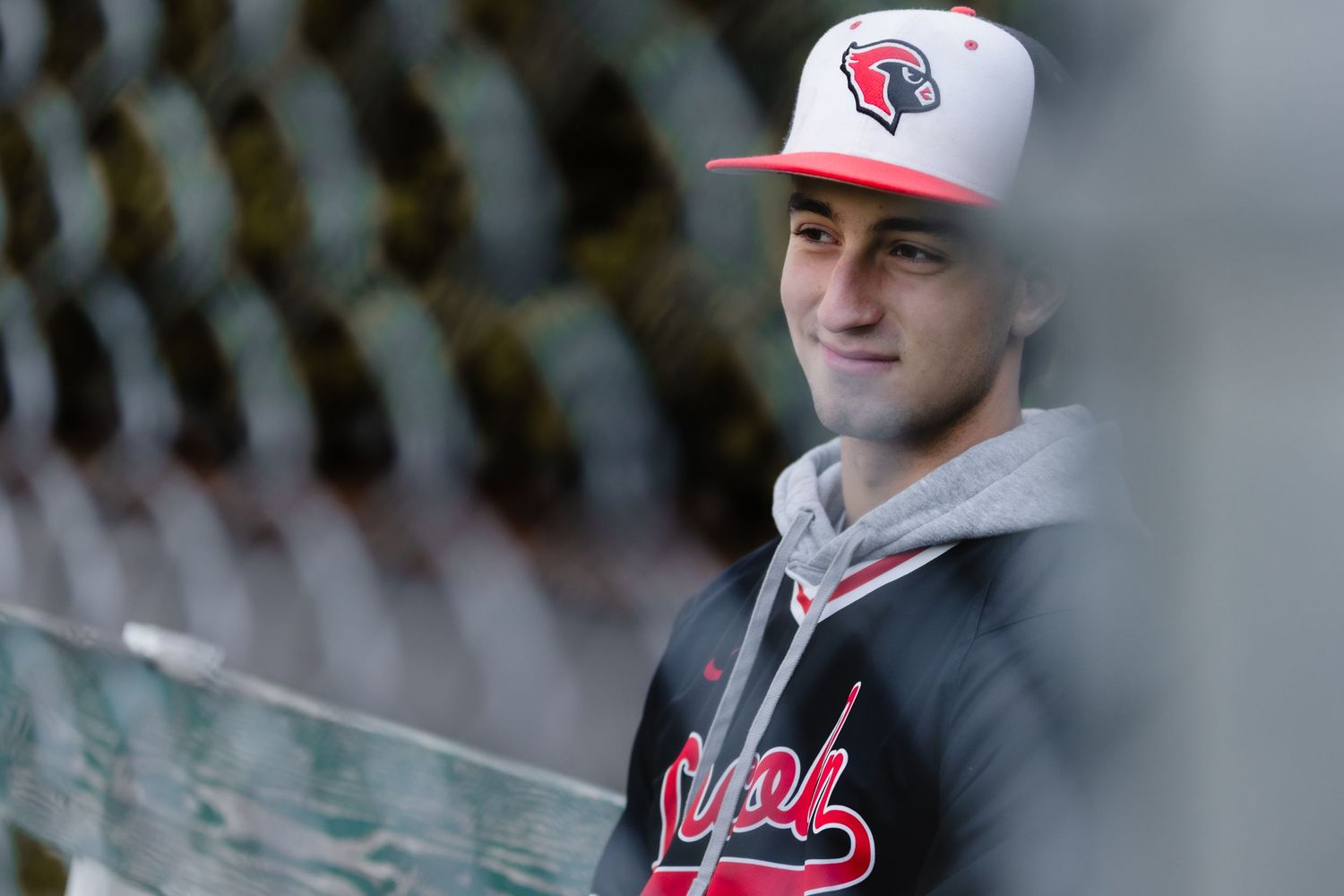 Baseball player wearing UNL team gear stands near dugout fence wearing white cap with red logo and gray sweatshirt.