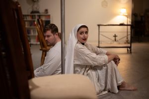 Two people in white robes sit back-to-back on the floor in a dimly lit room with warm lighting and minimal decor.